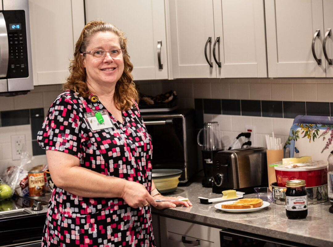 A lady in the kitchen at Pappus House smiling at the camera and about to butter some toast.