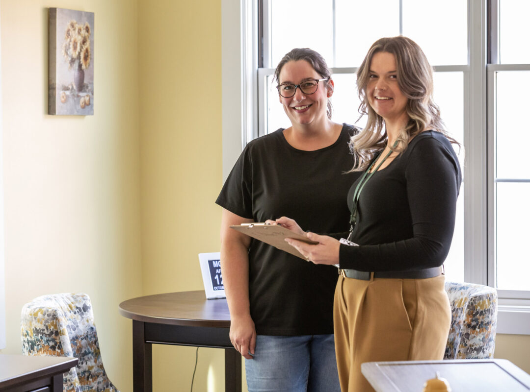 Two people smiling and looking at the camera. One of them is holding a clip board.