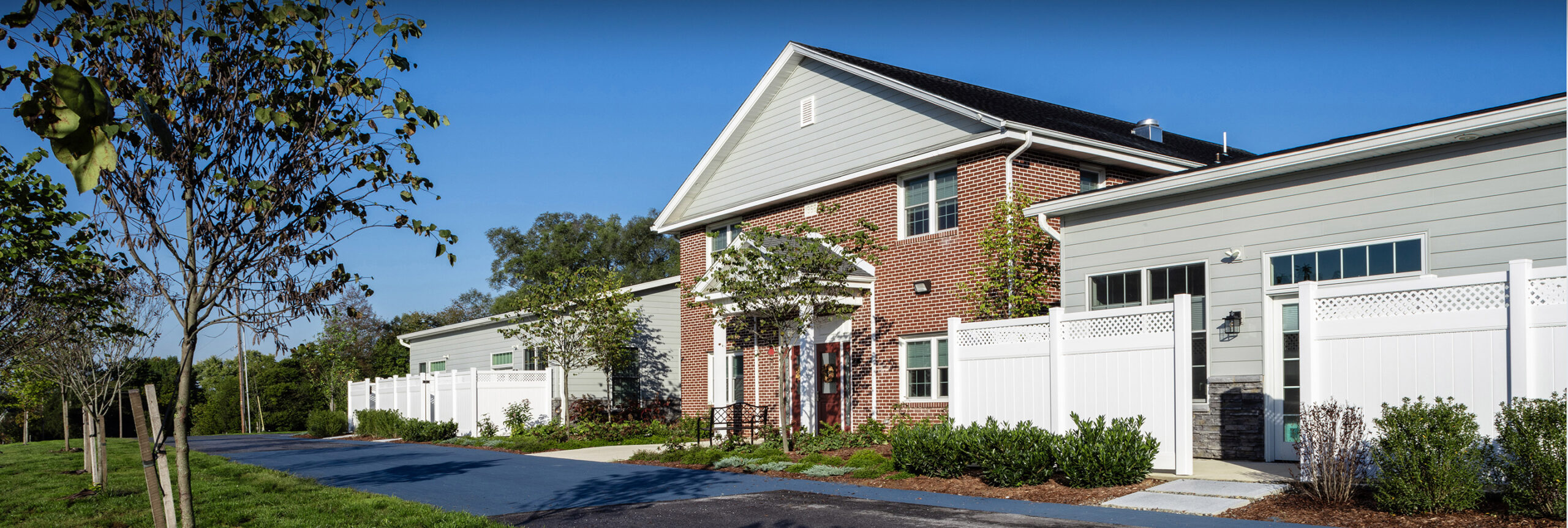 A house from the front verge with grass, bushes and a white fence around the front of the house.