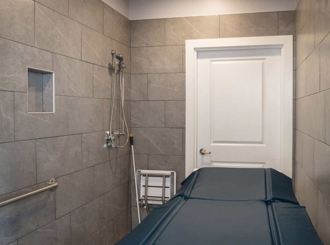 A tiled bathroom with small tiles on the floor and gray wall tiles. There is a bed in there to use for showering.