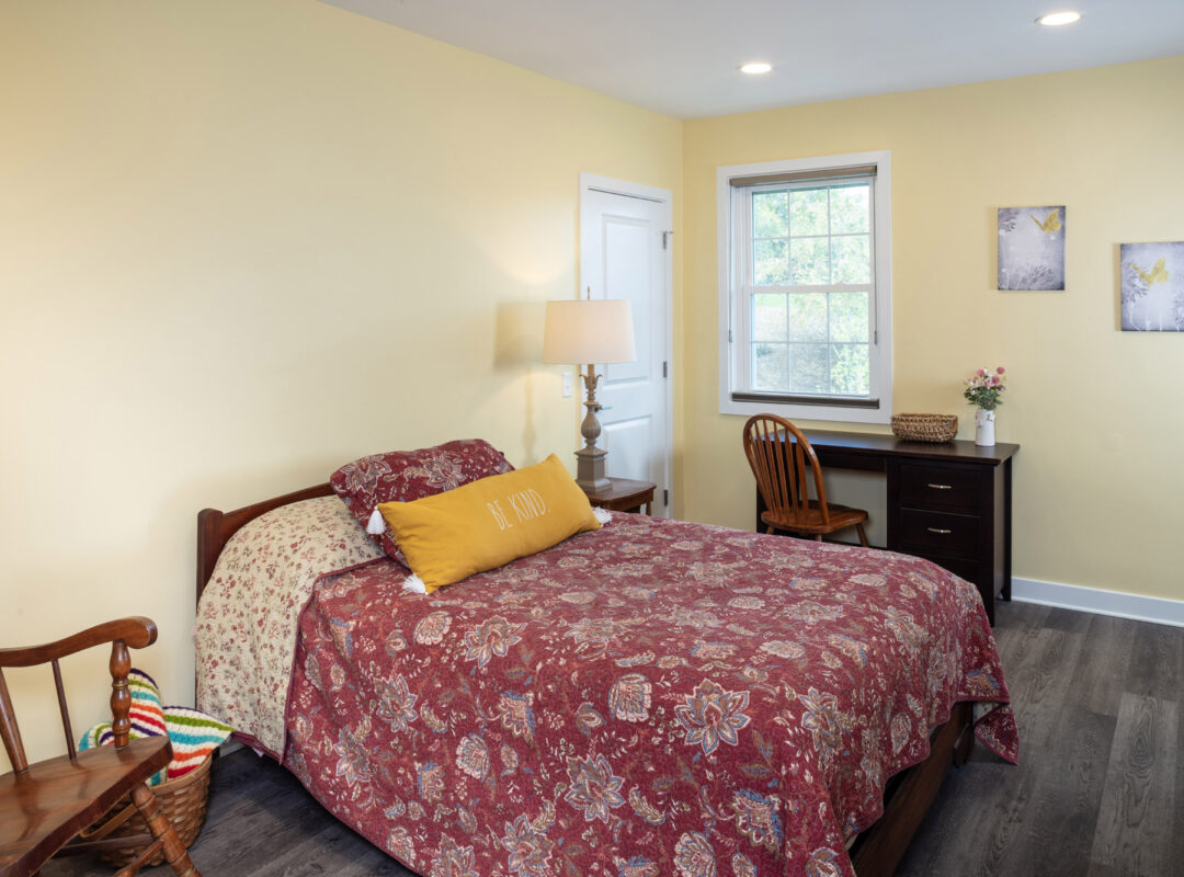 A yellow painted bedroom in Pappus House with a small desk, a rocking chair and a lamp next to the bed.